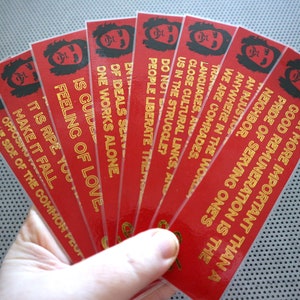 May include: A set of eight red bookmarks with gold lettering. Each bookmark features a black and white illustration of a person with a beard and afro. The text on the bookmarks reads: "Far more important than a good remuneration is the neighbor. Pride of serving one's..." and continues with different phrases on each bookmark.