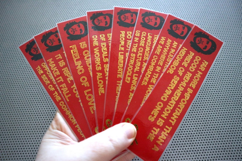 May include: A set of eight red bookmarks with gold lettering. Each bookmark features a black and white illustration of a person with a beard and afro. The text on the bookmarks reads: "Far more important than a good remuneration is the neighbor. Pride of serving one's..." and continues with different phrases on each bookmark.
