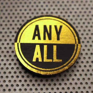 Puede incluir: Un botón redondo negro y dorado con las palabras "ANY ALL" escritas en letras doradas.