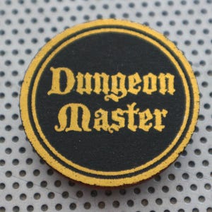 May include: A round black and gold Dungeon Master token. The text "Dungeon Master" is in gold lettering within a black circle, surrounded by a gold border. The token is on a gray, perforated surface.