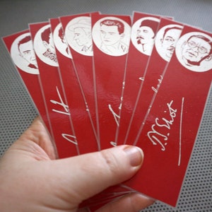 May include: A hand holds a fan of red bookmarks. Each bookmark features a white portrait of a man in a circle at the top. White script is printed on the red background. The bookmarks are glossy.