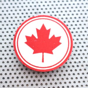 Op de afbeelding: Een rood esdoornblad op een witte cirkel met een rode rand. De cirkel bevindt zich op een zilveren achtergrond met een patroon van kleine gaten.