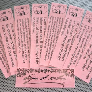 Op de afbeelding: Een verzameling roze papieren bladwijzers met gedrukte tekst en een geschetst gezicht. De bladwijzers zijn in een waaier gerangschikt, met een rechthoekig stuk onderaan met een handtekening en decoratieve rand.