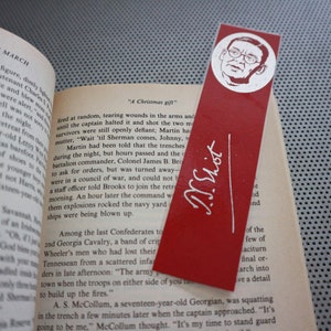 May include: Red bookmark with a white illustration of a man's face and the signature "T.S. Eliot" in white. The bookmark is placed in an open book with text visible.