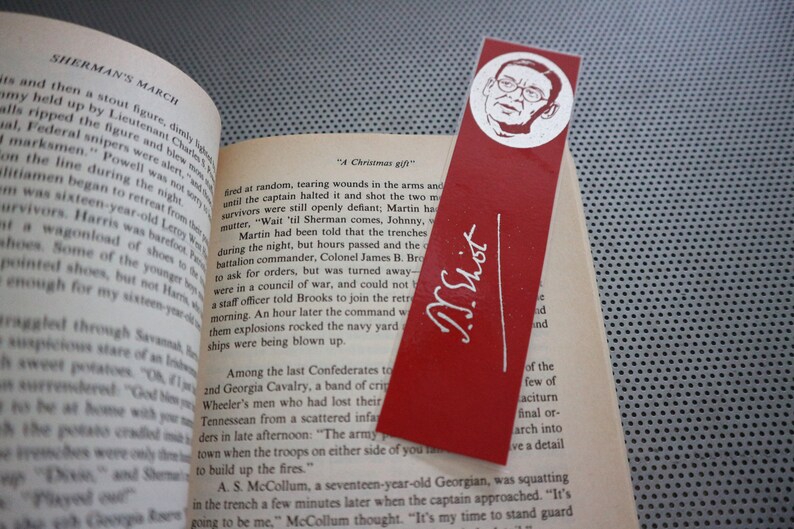 May include: Red bookmark with a white illustration of a man's face and the signature "T.S. Eliot" in white. The bookmark is placed in an open book with text visible.