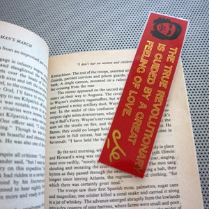 May include: A red bookmark with gold lettering that reads "The True Revolutionary is Guided by a Great Feeling of Love." The bookmark is placed on a page of a book about Sherman's March.
