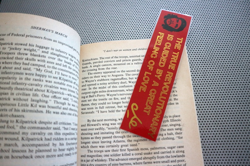 May include: A red bookmark with gold lettering that reads "The True Revolutionary is Guided by a Great Feeling of Love." The bookmark is placed on a page of a book about Sherman's March.