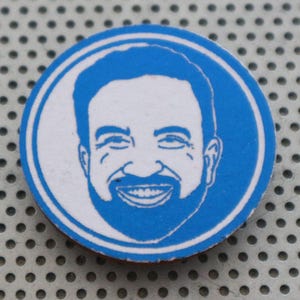 Könnte beinhalten: Ein runder, blau-weißer Knopf mit einem lächelnden Mann mit Bart. Das Gesicht des Mannes befindet sich in der Mitte des Knopfes, umgeben von einem blauen Rand. Der Hintergrund ist eine perforierte Metalloberfläche.