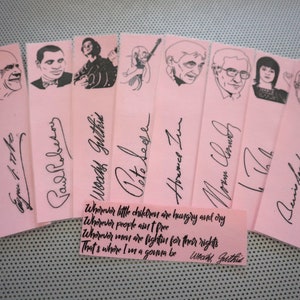 May include: A set of pink bookmarks with black ink portraits and signatures. A small pink rectangle displays a quote by Woody Guthrie. The bookmarks are arranged on a light blue surface.