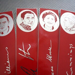 May include: Red bookmarks with silver portraits of notable figures. Each bookmark features a different person in a circular frame, with their name and signature in silver script below. The bookmarks are arranged in a row.