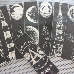 Może przedstawiać: Zestaw ośmiu czarno-białych naklejek z obrazkami i tekstem związanym z lądowaniem na Księżycu misji Apollo 11. Naklejki przedstawiają obrazy statku kosmicznego Apollo 11, Księżyca i astronautów Neila Armstronga i Buzza Aldrina. Tekst na naklejkach zawiera cytaty astronautów, takie jak "To mały krok dla człowieka, ale gigantyczny skok dla ludzkości." i "Tutaj ludzie z Ziemi po raz pierwszy postawili stopę na Księżycu."