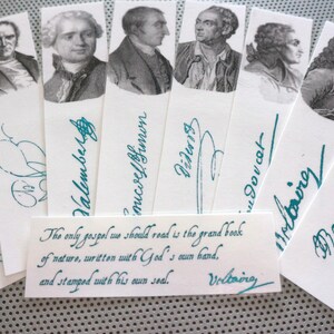 Puede incluir: Un conjunto de marcadores blancos con retratos de figuras históricas y firmas en color turquesa. También se incluye una pequeña tarjeta con una cita de Voltaire. Los marcadores están dispuestos en forma de abanico.
