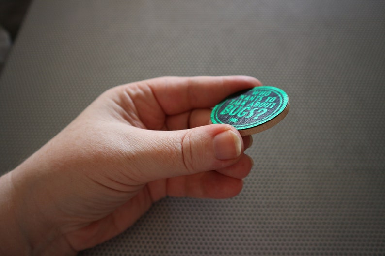 May include: A round wooden token with a black background and green text that reads "Who wants to talk about bugs?"
