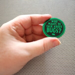 May include: A green and black round button with the text "WHO WANTS TO TALK ABOUT BUGS?" in white letters.