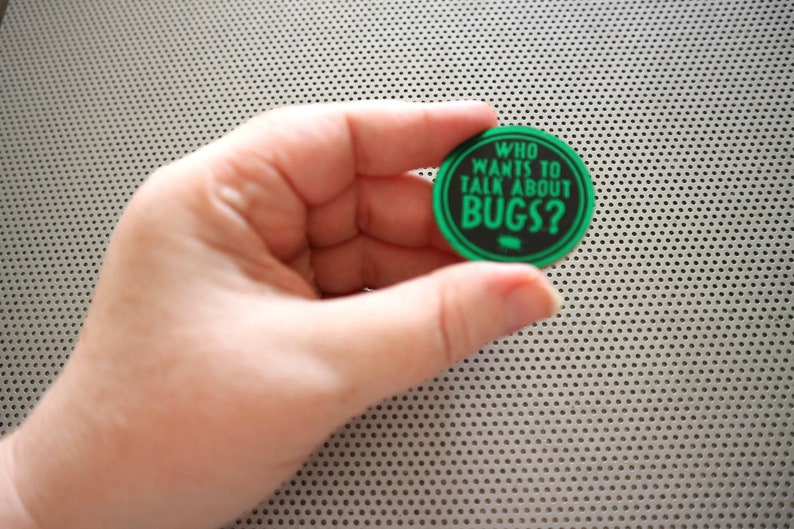 May include: A green and black round button with the text "WHO WANTS TO TALK ABOUT BUGS?" in white letters.