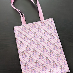 Peut inclure: Un sac fourre-tout rose avec un motif répété de dragons violets sur un fond floral rose clair. Le sac a des poignées roses et est fait de tissu.