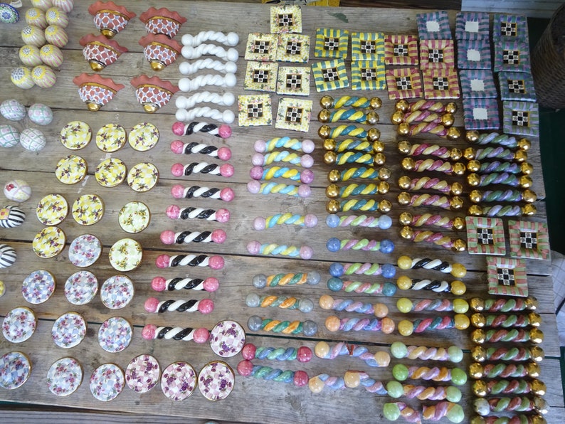 Puede incluir: Una colecci&oacute;n de pomos y tiradores decorativos de cer&aacute;mica en varios dise&ntilde;os y colores. El surtido incluye patrones florales, a rayas y geom&eacute;tricos, con detalles dorados y plateados. Los pomos y tiradores est&aacute;n dispuestos sobre una superficie de madera.