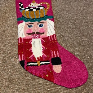 Może przedstawiać: Różowa świąteczna skarpeta z motywem postaci łamacza orzechów. Łamacz orzechów ma na sobie czerwony i biały kapelusz z czajnikiem na górze. Skarpeta jest ozdobiona złotymi i czarnymi akcentami.