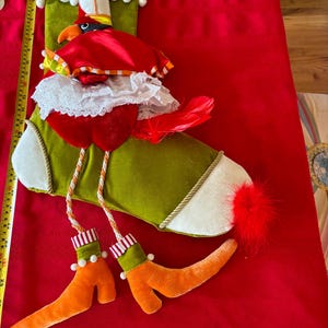 Puede incluir: Una media verde y blanca con un cardenal rojo que lleva un vestido de encaje blanco y un sombrero rojo. La media tiene un puño blanco con pompones blancos y piernas a rayas naranja y blancas.