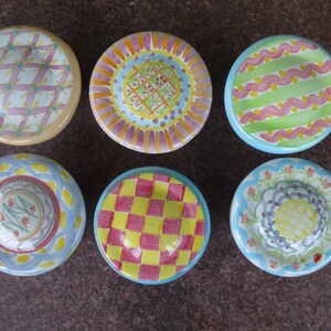 May include: Six small, hand-painted ceramic dishes with colourful, patterned designs. Each dish features a unique combination of colours and patterns, including checks, floral motifs, and geometric shapes. The dishes are arranged in a circular pattern.