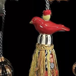 May include: A red bird finial with black and white stripes, gold trim, and a tassel made of yellow, red, and white threads.