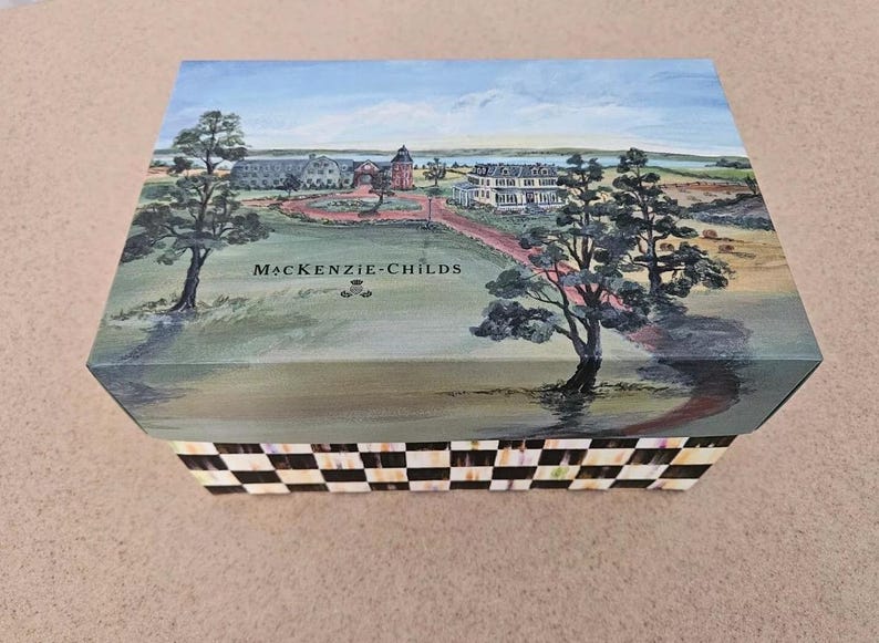 Op de afbeelding: Rechthoekige decoratieve doos met een geschilderde landschapssc&egrave;ne op het deksel. De sc&egrave;ne toont gebouwen, bomen en een pad, met de tekst "MacKenzie-Childs" op de voorkant. De zijkanten van de doos hebben een zwart-wit geblokt patroon.