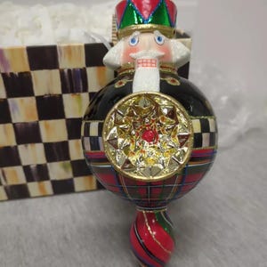 May include: A decorative Christmas ornament shaped like a nutcracker soldier. The ornament features a red, green, and gold crown, a white beard, and a black and plaid body. A gold starburst design is on the front, and the bottom is a red and green spiral.