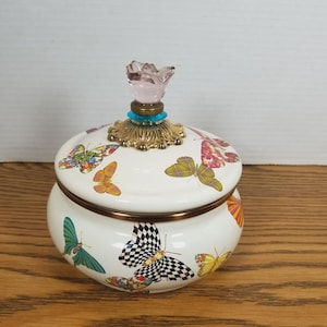 以下が含まれることがあります： カラフルな蝶のイラストが施された、白い装飾用の小物入れ。 蓋にはピンクの花の形をしたノブと金色のアクセントが付いています。 ボックスの直径は約10cmです。