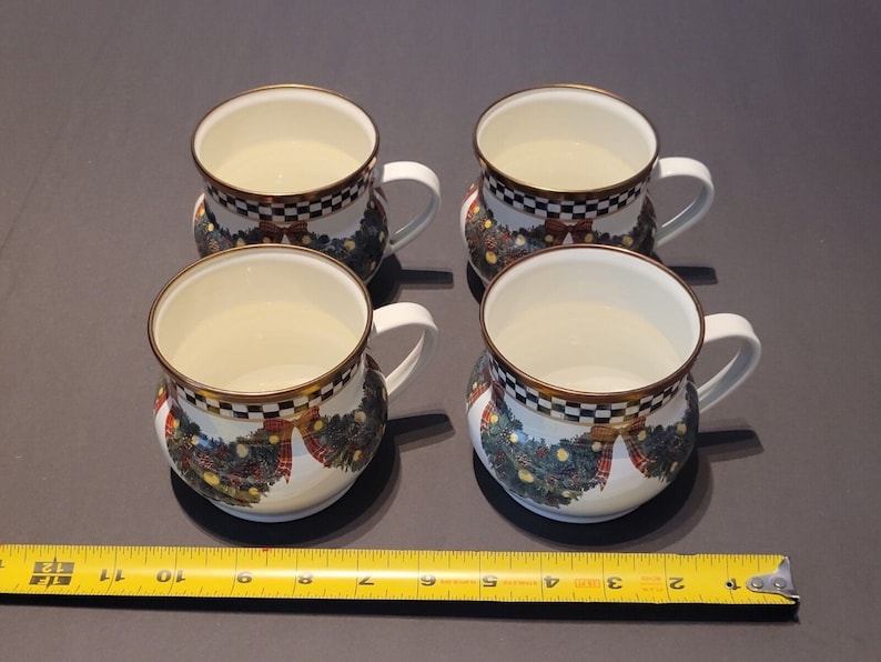 K&ouml;nnte beinhalten: Vier wei&szlig;e Keramikbecher mit goldenen R&auml;ndern und wei&szlig;en Griffen. Jeder Becher hat ein schwarz-wei&szlig; kariertes Band oben und ein festliches Kranzdesign mit roten Schleifen und gr&uuml;nem Laub. Die Becher haben einen Durchmesser von etwa 10 cm.