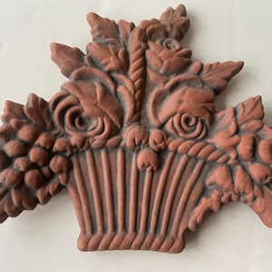 May include: A terracotta wall plaque depicting a basket overflowing with flowers and leaves. The basket is woven and has a rope handle. The flowers are stylized and have a raised texture.