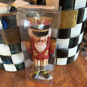 May include: A red, white, and gold nutcracker ornament with a black hat and a white beard. The ornament is in a clear plastic package with a gold string.