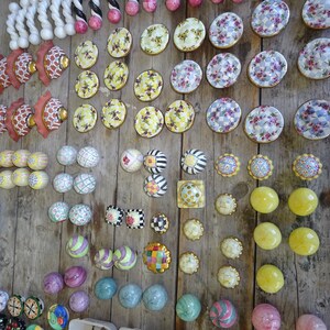 Puede incluir: Una colecci&oacute;n de pomos y tiradores decorativos para armarios en varios dise&ntilde;os y colores, incluyendo patrones florales, a rayas y geom&eacute;tricos. Los pomos son de cer&aacute;mica y metal, y est&aacute;n dispuestos sobre una superficie de madera r&uacute;stica.