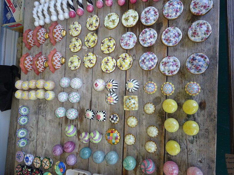 Puede incluir: Una colecci&oacute;n de pomos y tiradores decorativos para armarios en varios dise&ntilde;os y colores, incluyendo patrones florales, a rayas y geom&eacute;tricos. Los pomos son de cer&aacute;mica y metal, y est&aacute;n dispuestos sobre una superficie de madera r&uacute;stica.