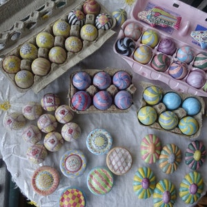 May include: A selection of decorative, hand-painted ceramic eggs and ornaments in various colours and patterns. The eggs are arranged in cardboard and pink egg boxes, and displayed on a white tablecloth. Some ornaments are flower-shaped.