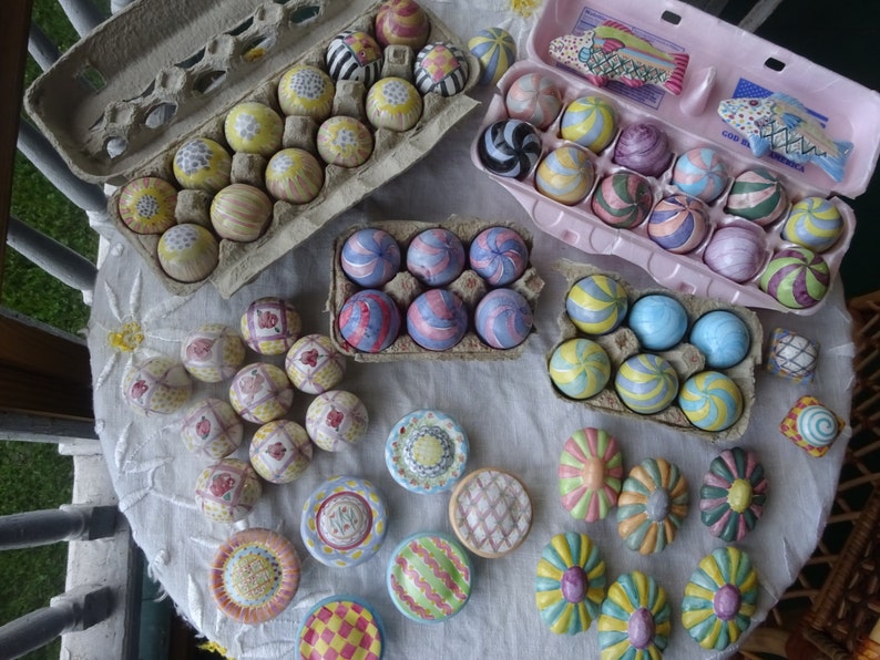 May include: A selection of decorative, hand-painted ceramic eggs and ornaments in various colours and patterns. The eggs are arranged in cardboard and pink egg boxes, and displayed on a white tablecloth. Some ornaments are flower-shaped.
