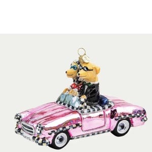 Op de afbeelding: Een roze cabrio-ornament met twee hondenfiguren die een zonnebril en formele kleding dragen. De auto heeft zwart-witte geruite accenten en het woord "Aurora" aan de voorkant.
