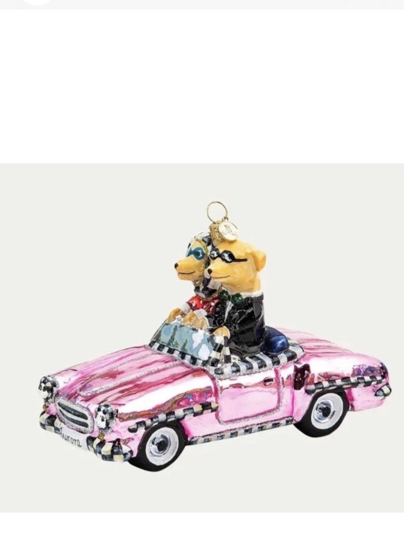 Op de afbeelding: Een roze cabrio-ornament met twee hondenfiguren die een zonnebril en formele kleding dragen. De auto heeft zwart-witte geruite accenten en het woord "Aurora" aan de voorkant.