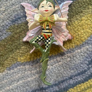 May include: A whimsical fairy ornament with pink wings, green hair, and a red and white striped outfit. The fairy is wearing a green and red bow tie and is standing on a green vine.