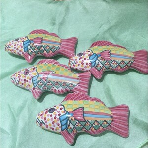 Puede incluir: Cuatro figuras de peces de cer&aacute;mica con patrones coloridos. Los peces tienen aletas y colas rosas, con cuerpos decorados con lunares y dise&ntilde;os geom&eacute;tricos en amarillo, azul y verde. Dispuestos sobre una superficie verde claro.