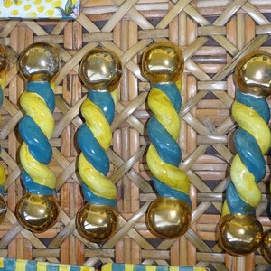 May include: Eight decorative cabinet pulls with a twisted design in yellow and teal. Each pull has gold-coloured spherical ends. The pulls are displayed against a woven background.