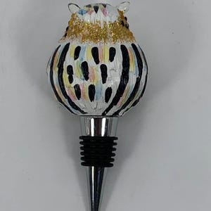 May include: A decorative bottle stopper featuring a whimsical owl design. The owl has a white body with black stripes and colorful accents. The head is adorned with gold glitter. The stopper has a silver metal base and a black rubber seal.