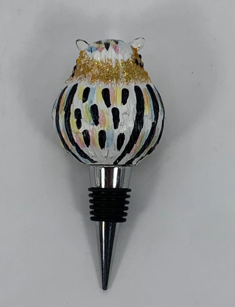 May include: A decorative bottle stopper featuring a whimsical owl design. The owl has a white body with black stripes and colorful accents. The head is adorned with gold glitter. The stopper has a silver metal base and a black rubber seal.