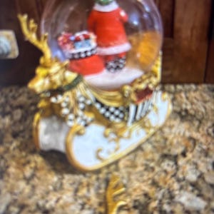 May include: A festive snow globe featuring a green character in a red coat and hat, standing on a snowy base. The globe sits atop a gold and white sleigh with reindeer details, ideal for holiday decor.