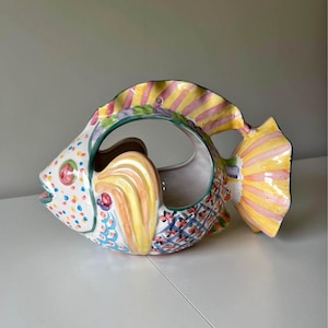 以下が含まれることがあります： 中央に吊り下げ用の穴が開いた、カラフルなセラミック製の魚のプランター。魚は黄色、ピンク、緑、青、白のアクセントで描かれています。