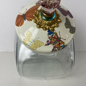 May include: A clear glass jar with a lid featuring a butterfly design. The lid has a pink heart-shaped knob, a gold-colored base, and a teal accent. The butterflies are in various colors, including yellow, orange, and blue.