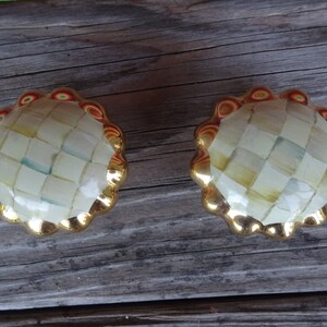 May include: A pair of decorative cabinet knobs. Each knob has a gold-coloured, scalloped edge and a checkered, off-white centre with hints of blue and yellow. Suitable for furniture and home decor.