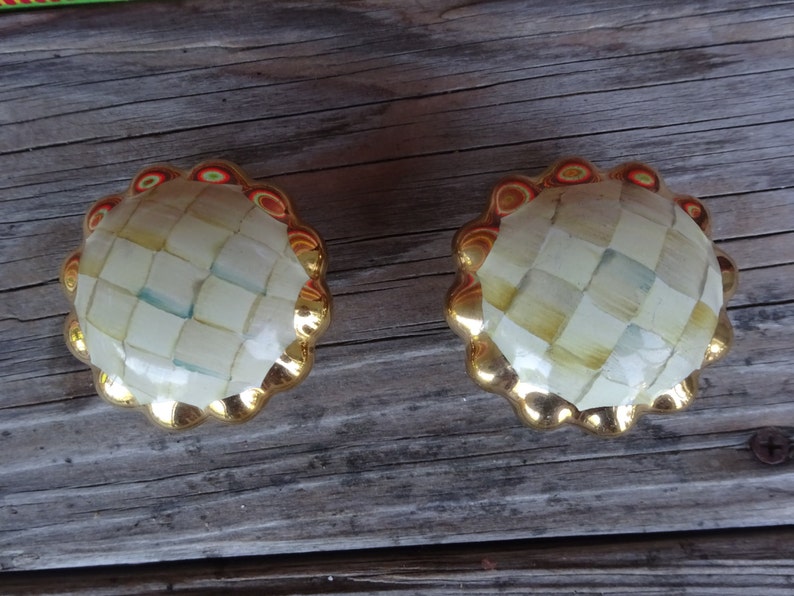 May include: A pair of decorative cabinet knobs. Each knob has a gold-coloured, scalloped edge and a checkered, off-white centre with hints of blue and yellow. Suitable for furniture and home decor.