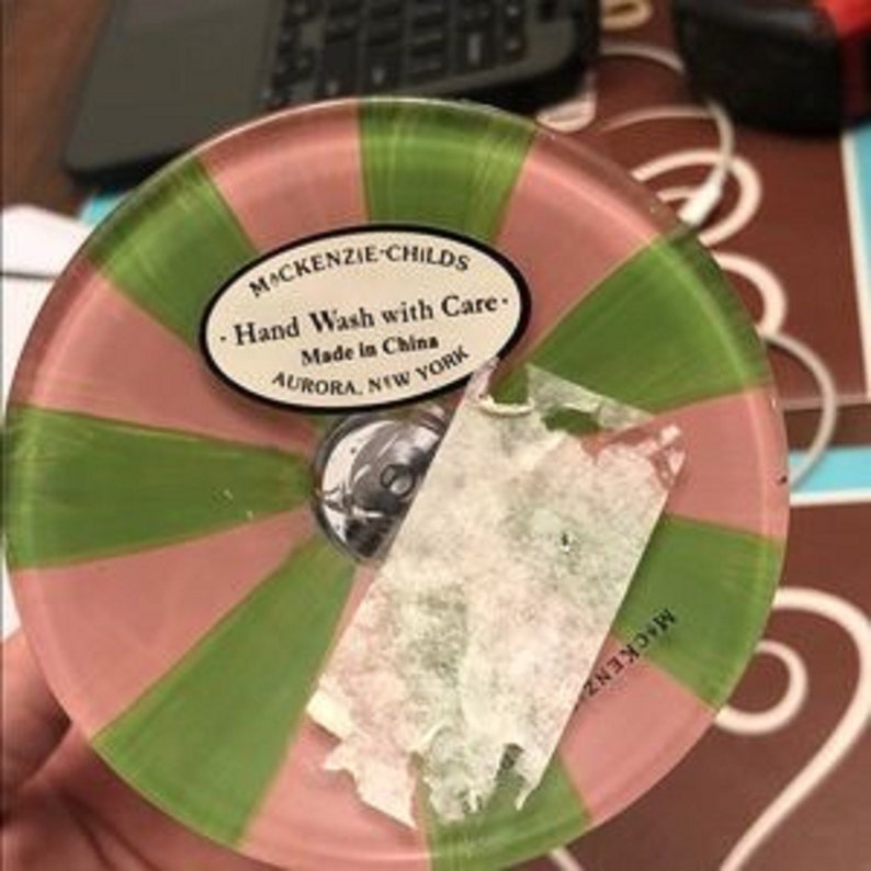 Puede incluir: Un posavasos decorativo de cristal con rayas rosas y verdes alternas. El posavasos tiene un centro de metal plateado y una etiqueta parcialmente despegada que dice "MACKENZIE-CHILDS Hand Wash with Care Made in China AURORA, NEW YORK."