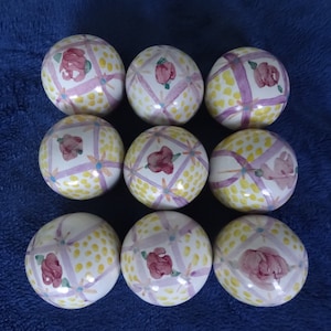May include: Nine decorative ceramic spheres with a white base, featuring a repeating pattern of yellow polka dots, lavender ribbons, and red rose floral designs. The spheres are arranged in a grid pattern against a blue background.