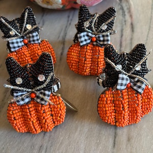 May include: Four beaded cat-shaped pumpkin decorations. Each cat has a black beaded head with silver accents and a gingham bow. The pumpkins are orange and beaded. A decorative pumpkin with floral patterns is in the background.